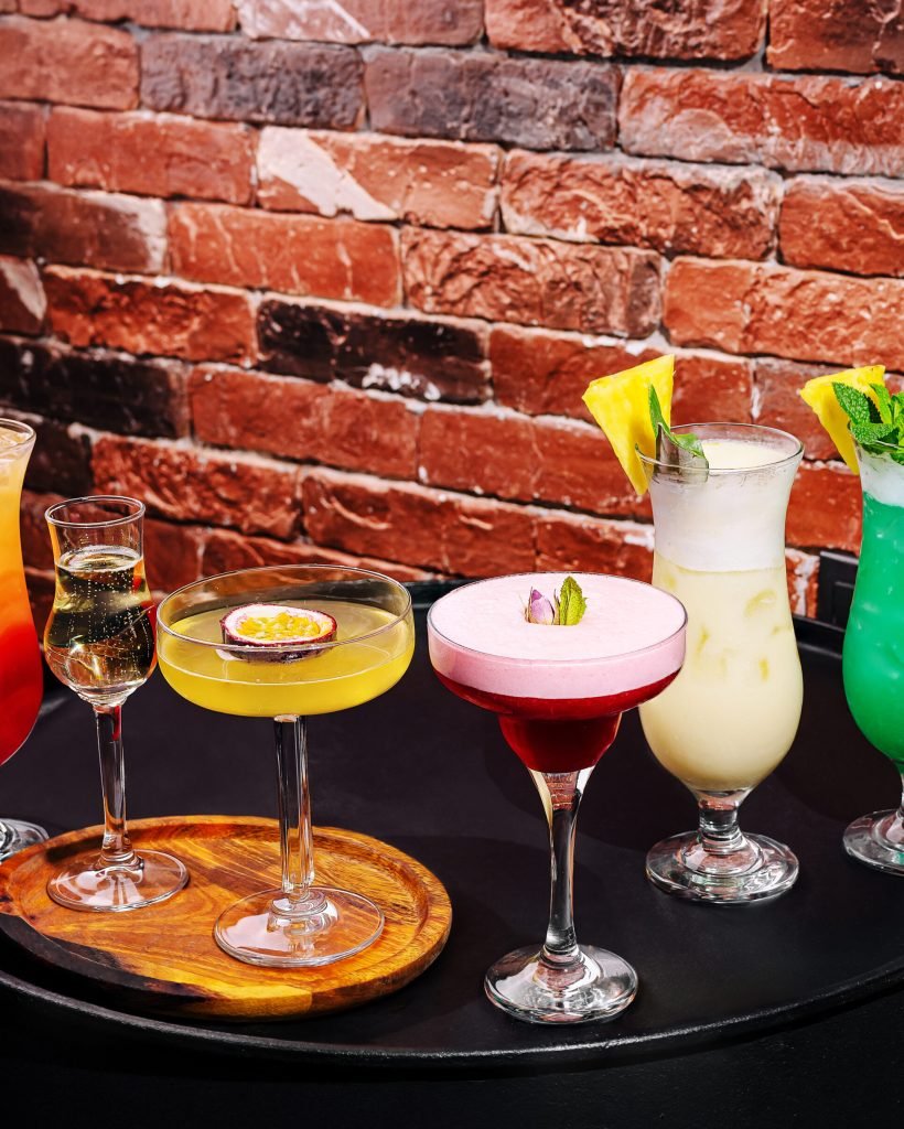 A variety of vibrant cocktails are displayed on a dark surface inside a bar. Each drink features unique garnishes, showcasing the creativity of mixology in an inviting atmosphere.